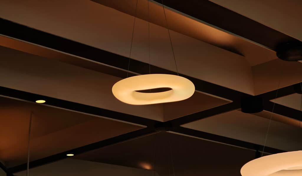 Lustre design torsadé jaune pâle suspendu sous un plafond moderne marron avec des poutres noires et des spots encastrés.