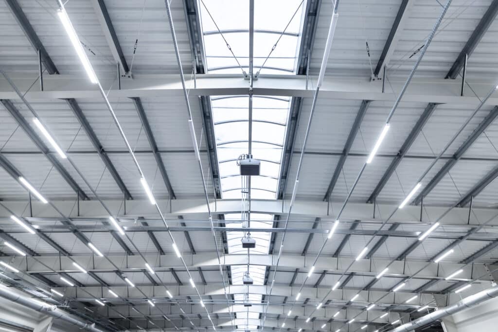 Intérieur d'un grand entrepôt industriel moderne avec toit métallique, puits de lumière zénithale et longs luminaires LED blancs.