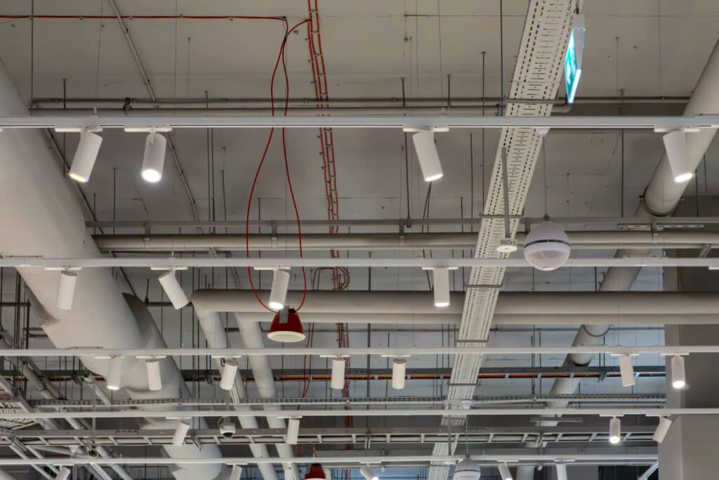 Plafond technique d'un commerce avec conduits d'aération blancs, chemins de câbles perforés, projecteurs orientables blancs et un panneau de sortie de secours vert.